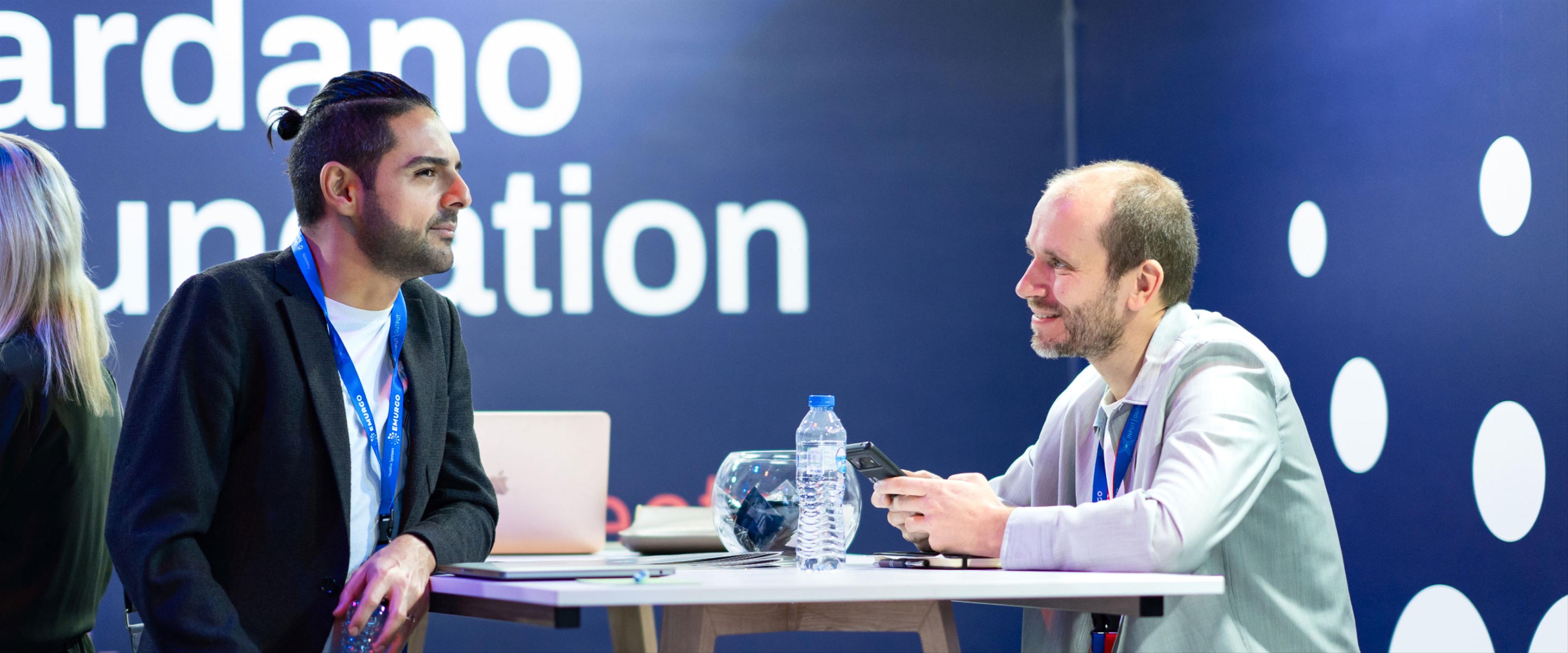 A photograph of Sebastian Bode engaged in a discussion at the Cardano Foundation booth. 